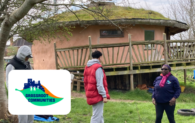 A small group of people stands outside a unique eco-friendly structure with a green, grass-covered roof and wooden balcony. The setting is surrounded by trees and grassy terrain, exuding a sense of community and sustainability. A logo for "Grassroot Communities" is placed in the corner of the image.